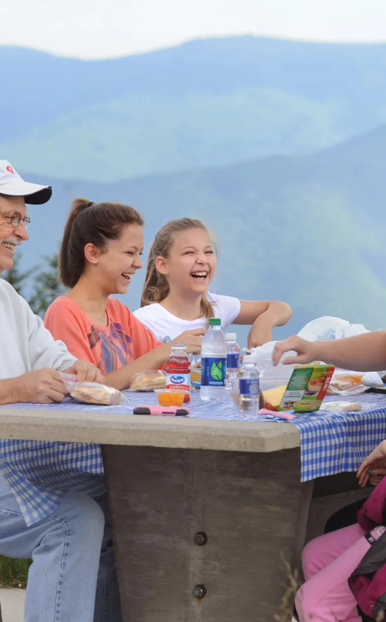Three adults of different generations sit at a picnic table in the mountains