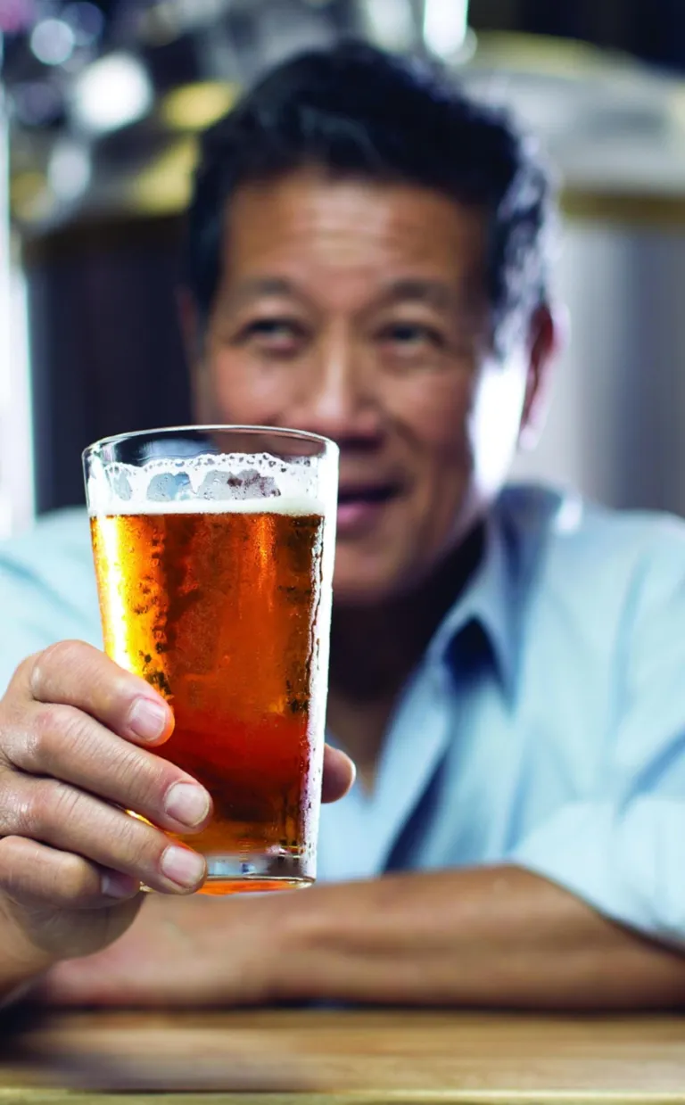 a person in a dress shirt smiles while holding a beer in front of them at a bar