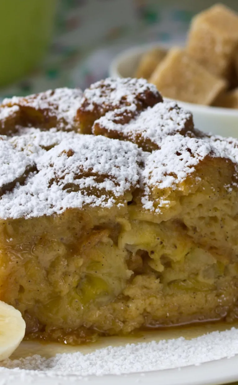 a square of banana bread pudding sprinkled with powdered sugar and sliced bananas on the side